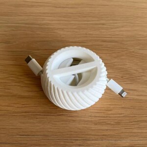 Op de afbeelding: Een witte kabelorganizer met een ronde vorm en een spiraalpatroon. Het heeft twee kabels, één met een USB-C-connector en één met een Lightning-connector. Dit product is ontworpen voor kabelbeheer en -opslag.
