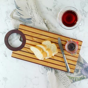 May include: A wooden serving board with a dark brown handle, slices of bread, a small jar of jam, and a knife. A glass of tea and a decorative cloth are also present. The board has alternating light and dark wood stripes.