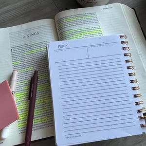 Könnte beinhalten: Eine offene Bibel mit markiertem Text und ein Spiralnotizbuch mit der Aufschrift "Prayer" liegen auf einer Holzoberfläche. Ein Stift, ein pinkfarbener Haftzettel und eine Tasse mit der Aufschrift "hello gorgeous" sind ebenfalls vorhanden, was auf eine Studien- oder Andachtsumgebung hindeutet.