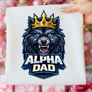 May include: White t-shirt with a wolf illustration wearing a gold crown, and the text "ALPHA DAD" in blue and white. The wolf has blue eyes and an aggressive expression. The design is bold and striking.