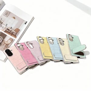 Puede incluir: Una colección de fundas para teléfonos de colores pastel, incluyendo rosa, morado, amarillo y azul. Cada funda presenta un patrón texturizado similar a la piel de cocodrilo y un marco plateado. Algunas fundas tienen un soporte incorporado.