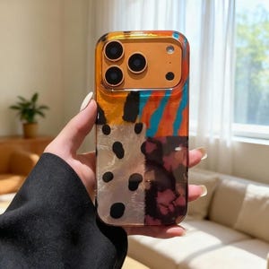 Può includere: Custodia trasparente per telefono con un colorato design astratto. La custodia presenta motivi arancioni, neri, bianchi e blu. La sezione della fotocamera è dorata e ha tre lenti.