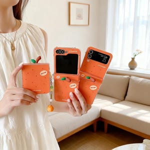 May include: Orange phone cases with a fruit design. The cases have a flip design and feature a small green leaf and a brown stem. The word "Orange" is printed on the cases. A small orange charm hangs from one of the cases.