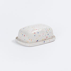 May include: A white ceramic butter dish with a lid, decorated with colorful paint splatters. The dish has a rectangular base and a rounded, domed lid. The paint splatters are in various colors, including blue, red, yellow, and green.