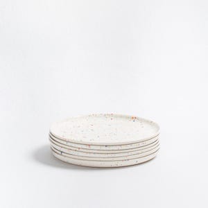 May include: A stack of six off-white ceramic plates with a speckled design. The plates have a simple, round shape and are decorated with small, colorful flecks of red, blue, and orange. The plates are stacked on top of each other.
