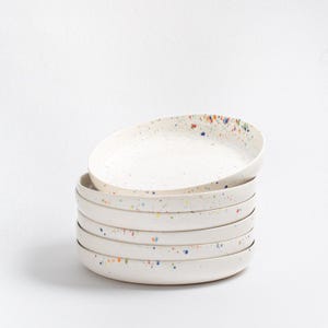 May include: A stack of six white ceramic plates with a speckled design. The plates have a slightly raised edge and are decorated with colorful paint splatters around the rim. The plates are stacked on top of each other.
