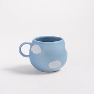 May include: A light blue ceramic mug with a rounded shape and a small handle. The mug features white cloud designs on the exterior. The mug is designed for beverages.