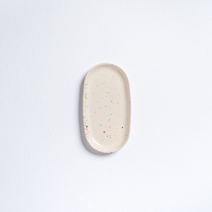 Peut inclure: Un plateau en céramique de forme ovale et de couleur crème, parsemé de mouchetures colorées. Le plateau a une surface lisse et un bord légèrement surélevé. Les mouchetures sont de différentes couleurs, dont le rouge, le bleu et le jaune. C'est un objet décoratif.