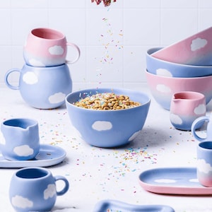 May include: A collection of blue and pink ceramic tableware with a cloud motif. The set includes mugs, bowls, and a small pitcher. Some bowls are filled with cereal, and sprinkles are falling from above.