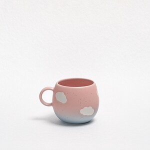 May include: A pink ceramic mug with a rounded shape and a small handle. The mug features white cloud designs and a gradient from pink to light blue at the base. The mug is designed for hot beverages.