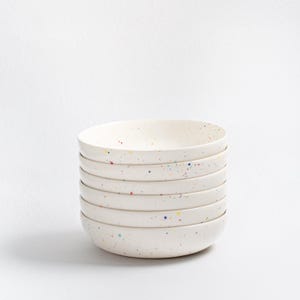 May include: A stack of seven off-white ceramic bowls. Each bowl features a speckled design with colourful dots in red, yellow, and blue. The bowls are stacked neatly, showcasing a simple, modern design, suitable for everyday use.