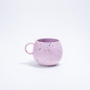 May include: A light purple ceramic mug with a rounded shape and a small handle. The mug is speckled with small, colorful dots in various colors. The mug is designed for hot beverages.