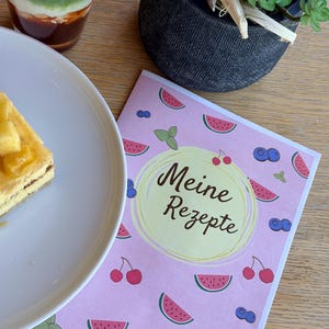 May include: A rectangular recipe book with a light pink cover featuring a repeating pattern of watermelon slices, cherries, blueberries, and mint leaves. A central yellow circle contains the black text Meine Rezepte in script font.