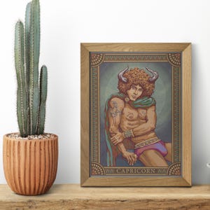 May include: A framed print of a Capricorn zodiac illustration. The artwork depicts a person with curly hair, horns, and a green cape. The print is in a wooden frame and is displayed on a wooden surface next to a cactus in a terracotta pot.