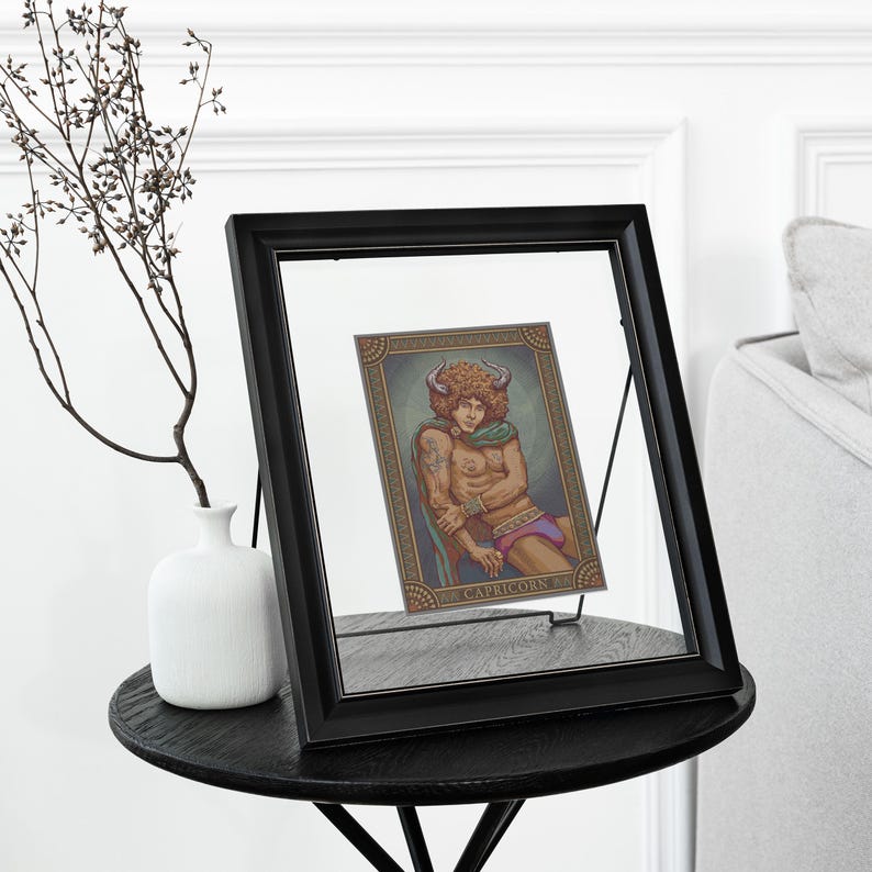 May include: A framed art print featuring a Capricorn zodiac illustration. The artwork depicts a figure with horns, a green scarf, and purple shorts. The black frame sits on a dark round table, with a white vase and decorative branch nearby.