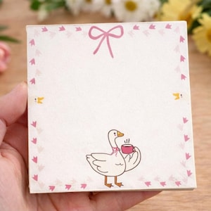 Puede incluir: Un bloc de notas cuadrado con un lazo rosa en la parte superior y un borde de corazones rosas. El bloc de notas presenta un ganso de dibujos animados sosteniendo una taza rosa. El bloc de notas se sostiene en una mano.