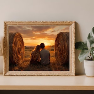 Op de afbeelding: Een ingelijste prent toont een stel dat naar een zonsondergang in een veld kijkt, met twee grote hooibalen aan weerszijden. Het kunstwerk zit in een goudkleurige lijst en rechts staat een plant in een pot.