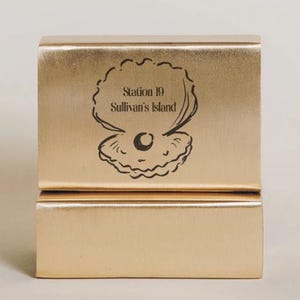 May include: A gold-colored, rectangular box with a hinged lid. The box features a black illustration of an oyster with a pearl and the text "Station 19 Sullivan's Island". The box is made of a shiny, metallic material.