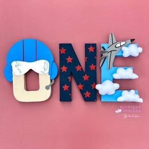 Könnte beinhalten: Ein Holzschild "ONE" mit Luftfahrt-Thema. Das "O" ist ein Pilothelm, das "N" hat rote Sterne auf marineblauem Hintergrund, und das "E" hat ein graues Flugzeug und weiße Wolkenakzente. Der Hintergrund ist rosa.