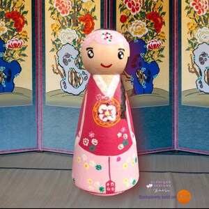 Puede incluir: Muñeca de madera pintada a mano con vestimenta tradicional coreana. La muñeca presenta un vestido rosa y rojo con detalles florales y un colgante decorativo. La cara de la muñeca tiene una expresión alegre, con una pantalla coreana tradicional.