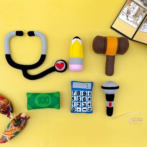 May include: A collection of felt toys including a stethoscope, pencil, gavel, microphone, calculator, and a $100 bill, arranged on a yellow surface. The stethoscope has a heart detail. The toys are designed with bright colors and simple shapes.