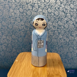 Puede incluir: Muñeca de madera pintada a mano que representa una figura con ropa tradicional coreana. La muñeca tiene una chaqueta azul claro, un cuello blanco y una falda gris. La cara está pintada con ojos negros y una sonrisa roja. La muñeca está sobre una superficie de madera.