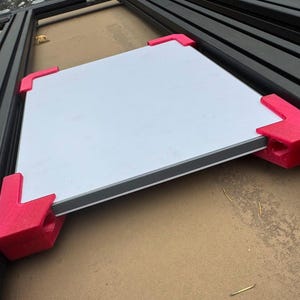 May include: A white, square panel with a gray border, secured by four bright pink corner brackets. The panel is resting on a light brown surface, with black metal framing visible in the background. The brackets appear to be 3D-printed.