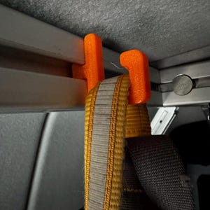 May include: Close-up of an orange 3D-printed hook attached to a metal frame. A yellow and gray strap is hanging from the hook. The hook is designed to hold items within a vehicle.