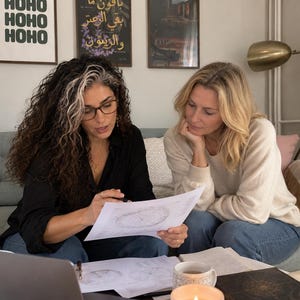 Lifestyle photograph of astrologer Selene sitting on a sofa beside a female client in a cozy living room. Wearing glasses and a black blouse, Selene uses a pen to explain a printed natal birth chart while the client listens closely. Astrology charts, a laptop and notes are spread across the coffee table. The scene illustrates a personal approach to 2027 astrology forecasts, 12 month year ahead readings, planetary transit analysis and birth chart predictions for love, career and life changes.