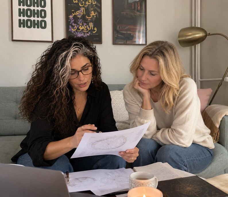 Lifestyle photograph of astrologer Selene sitting on a sofa beside a female client in a cozy living room. Wearing glasses and a black blouse, Selene uses a pen to explain a printed natal birth chart while the client listens closely. Astrology charts, a laptop and notes are spread across the coffee table. The scene illustrates a personal approach to 2027 astrology forecasts, 12 month year ahead readings, planetary transit analysis and birth chart predictions for love, career and life changes.