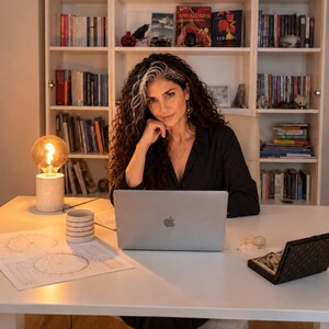 Selene, an experienced intuitive reader, sits at her desk in a cozy home office, surrounded by bookshelves, with a laptop, printed birth charts, warm glowing lamp and notebook. This behind-the-scenes photo shows the personal setting where she prepares individualized 2027 astrology forecasts and 12 month year ahead readings. Each birth chart forecast explores planetary transits, love and relationships, career, money, personal growth and important timing windows, delivered as a PDF report.