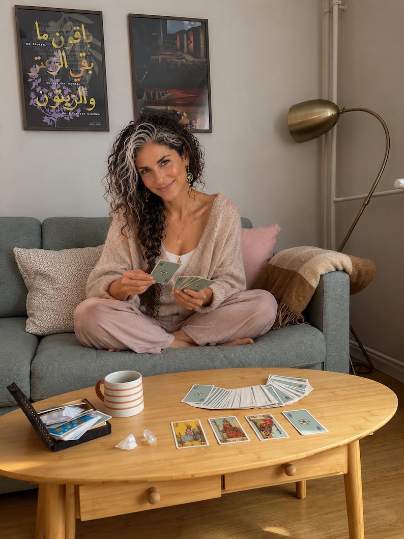 Selene, an intuitive psychic and tarot reader, sits comfortably on a sofa in her cozy living room, holding tarot cards beside a wooden coffee table with a tarot spread, crystals and a mug. This lifestyle photo introduces the welcoming atmosphere of AstroSoulHouse, offering love tarot readings, relationship guidance, psychic predictions, thoughts and feelings readings, and personalized spiritual insight. Explore her 2027 astrology forecast, birth chart reading and 12 month year ahead report.