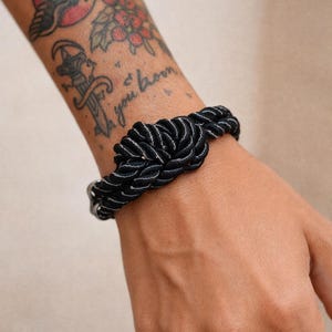 May include: Black and white rope bracelet with a nautical knot design. The bracelet is made of a thick, woven rope and features a silver-coloured clasp. A stylish accessory for everyday wear.