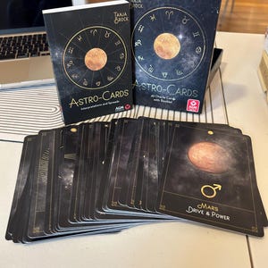 May include: Astro-Cards deck by Tanja Brock. The box and cards feature a black background with gold astrological symbols and a central golden orb. The cards are spread out, with one showing the planet Mars and the words "Mars Drive & Power."