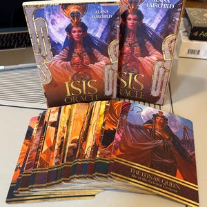 Könnte beinhalten: Ein Kartenspiel "Isis Oracle" von Alana Fairchild. Die Kartenbox zeigt eine Frau mit goldenem Kopfschmuck. Die Karten sind aufgefächert, eine zeigt "The Lunar Queen". Die Farbpalette umfasst Gold, Orange und Blau.
