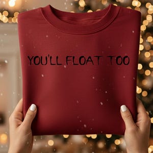 Könnte beinhalten: Ein weinrotes Sweatshirt mit dem Text "YOU'LL FLOAT TOO" in Schwarz. Das Sweatshirt wird vor einem Weihnachtsbaum mit Lichtern und einem Kamin hochgehalten. Schneeflocken fallen im Hintergrund.