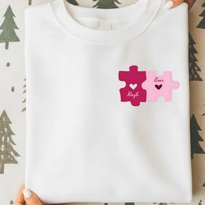 Pode incluir: Camisola branca com um design de peças de quebra-cabeça em tons de rosa. As peças do quebra-cabeça conectadas exibem os nomes "Nazli" e "Enes", e símbolos de coração. A camisola é segurada por uma pessoa.