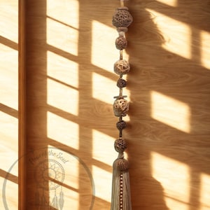 May include: A decorative wall hanging featuring a series of woven spheres and beads, culminating in a tassel. The design incorporates natural tones of beige and brown, with a metal ring at the top for hanging. The piece is displayed against a wooden background.