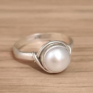 Könnte beinhalten: Ein silberner Ring mit einer großen, runden, weißen Perle als Mittelpunkt. Die Perle ist in einer Lünettenfassung gefasst, und das Band hat ein Design aus gedrehtem Draht. Der Ring wird auf einer hellbraunen Holzoberfläche gezeigt.