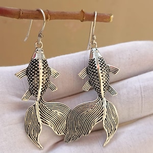 Könnte beinhalten: Ein Paar silberfarbene Fischohrringe. Jeder Ohrring hat ein detailliertes Fischdesign mit strukturierten Schuppen und fließenden Schwanzflossen. Die Ohrringe hängen an einfachen Ohrhaken und zeigen ein nautisches oder aquatisches Thema.