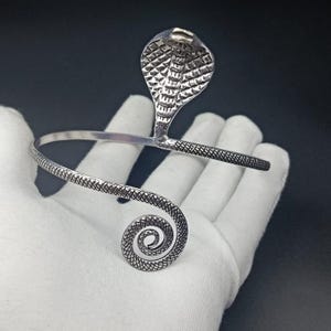 May include: A silver-toned arm cuff featuring a cobra design. The cuff has a coiled snake tail and a cobra head with detailed scales. The arm cuff is designed to be worn on the upper arm.