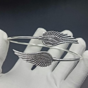 Puede incluir: Una pulsera de brazalete plateada con dos diseños detallados de alas. Las alas están situadas a ambos lados de la pulsera, con intrincados detalles de plumas. La pulsera se muestra sobre un fondo oscuro.