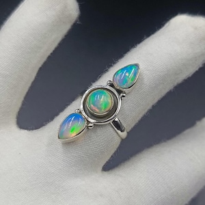 May include: A silver ring featuring three iridescent opals. The center stone is round and bezel-set within a circular frame, while the two flanking stones are pear-shaped. Each opal displays flashes of green, blue, and orange color play.