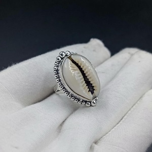 Puede incluir: Un anillo de plata con una concha de cauri blanca de forma ovalada como pieza central. La concha está engastada en un marco de plata decorativo adornado con un borde de cuentas. El anillo de metal tiene un acabado pulido y texturizado.