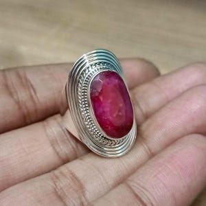 Puede incluir: Un anillo de plata con una gema grande, ovalada y de color rojo intenso. El anillo tiene un diseño detallado y texturizado alrededor de la gema, con múltiples líneas concéntricas y un borde trenzado. El anillo está sobre una mano.