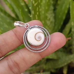 Puede incluir: Colgante de plata con un centro blanco en forma de espiral y un borde plateado con forma de cuerda. El colgante es redondo y tiene un pequeño lazo en la parte superior para una cadena. El fondo es verde borroso.