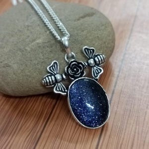 May include: Silver-toned pendant necklace featuring a dark blue, oval-shaped stone with sparkling inclusions. The pendant is adorned with two bee-shaped charms and a rose. The necklace is displayed on a stone surface.