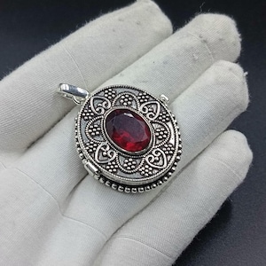 Könnte beinhalten: Ein ovaler silberfarbener Medaillon-Anhänger mit einem geprägten Muster aus kleinen Perlen und herzförmigen Schnörkeln. Ein facettierter, tiefroter ovaler Edelstein ist mittig in den Metallrahmen eingefasst, der eine kleine Öse besitzt.