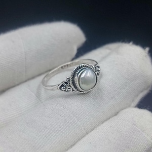 Op de afbeelding: Een zilveren ring met een centrale ronde witte parel gezet in een gedraaide touwrand. De band vertoont ingewikkelde krul- en filigraandetails aan beide zijden van de zetting, wat zorgt voor een decoratieve afwerking van het metaalwerk.