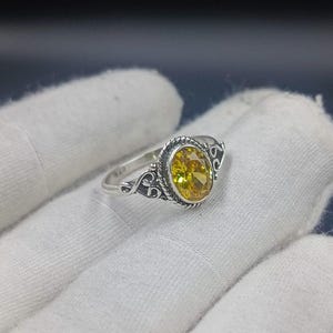 Può includere: Un anello in argento con una pietra preziosa gialla dal taglio ovale incastonata in una lunetta a corda intrecciata. La fascia presenta dettagli a volute su entrambi i lati e l'interno è chiaramente marchiato con il numero 925 per l'argento.